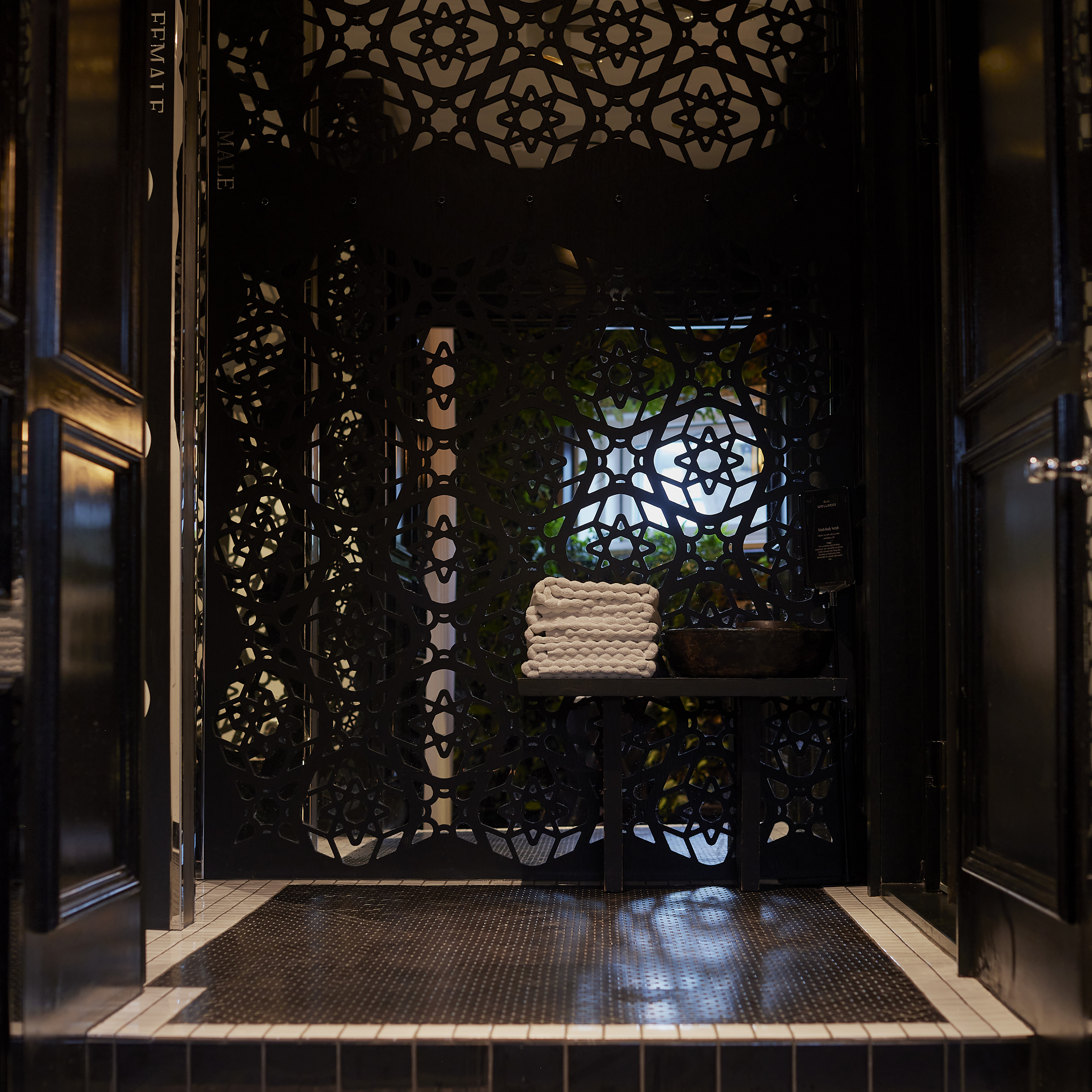 Dimly lit room with dark, ornate patterns on the walls and doors, and a stack of white towels on a black table.