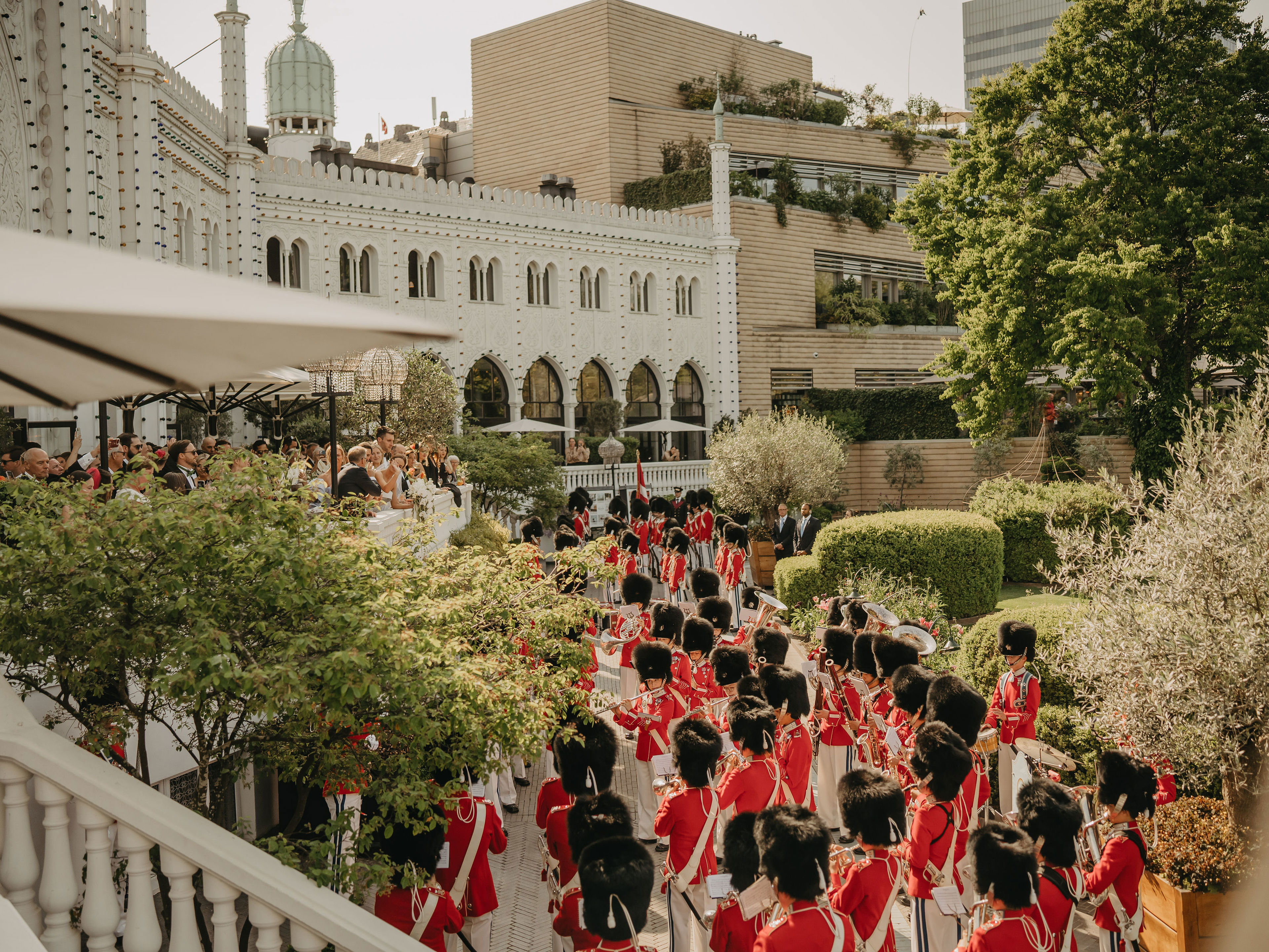Guards in red uniforms and bearskin hats march through garden near ornate building, crowd watching.